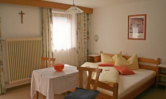 A simple room with a bed, a table, and chairs. There is a window with curtains and decorative cushions on the bed.