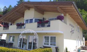A light yellow building with wooden cladding and balcony flowers. In front of the house is a curved entrance arch.