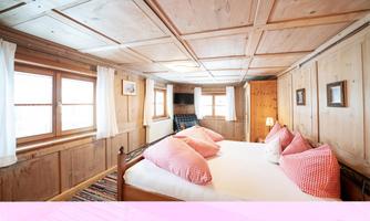 A cozy bedroom with wooden walls and large windows. The bed is decorated with pink pillows, and the room exudes rustic charm.
