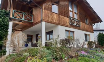 A beautiful wooden house with a green garden. It has large windows and balconies, surrounded by colorful flowers.