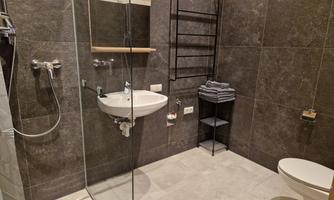 A modern bathroom with gray tiles and a glass shower. It includes a sink, a toilet, and a towel rack.
