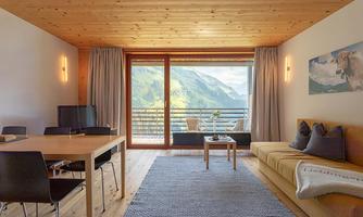 A modern living room with wooden details and a view of the mountains. There is a dining table, a cozy sofa area, and large windows that let in plenty of light.