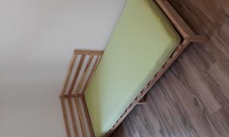 A simple wooden bed with a green mattress is in a room. The wall is light and there is a corner with green curtains.