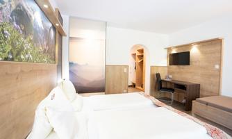 A modern hotel room with two single beds and a wood décor wall. There is a desk, a television, and a beautiful view of the mountains.