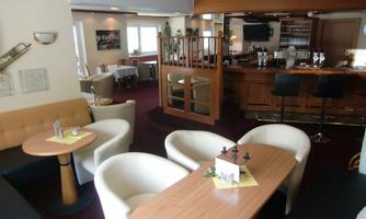 A cozy lounge with comfortable armchairs and wooden tables. In the background, there is a bar with stools.
