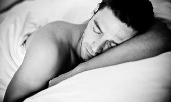 A man sleeps peacefully in a bed. He is lying on his side and has his eyes closed.