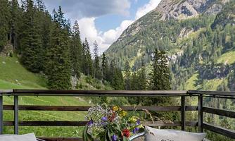 A cozy terrace with a table and chairs offers a view of the green mountains and the blue sky. A bouquet in a vase complements the inviting atmosphere.