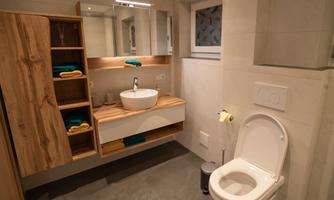 A modern bathroom with a sink, a mirror, and a toilet. Wooden shelves provide storage space for towels and toiletries.