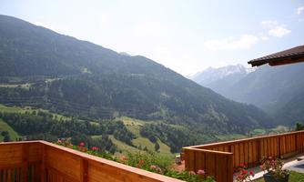 A picturesque mountain landscape with green hills and high mountains in the background. In the foreground, a wooden terrace with flowers.