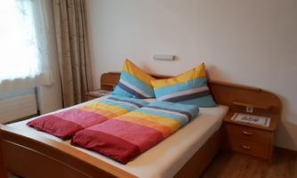 A cozy bedroom with a large double bed and colorful cushions. Next to the bed, there is a bedside lamp and a table.