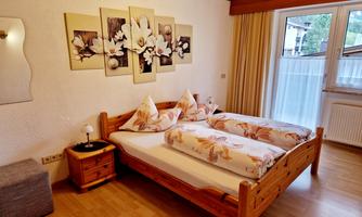 A cozy bedroom with a wooden double bed and decorative cushions. Pictures of flowers hang on the wall, and a window provides daylight.