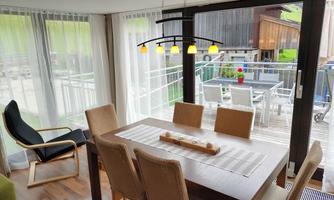 A modern dining room with a wooden table and chairs. Large windows offer a view of the terrace and the garden.