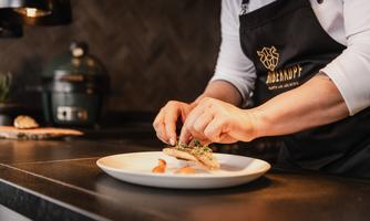 A chef arranges a dish on a white plate. The kitchen area is modern and well-lit.