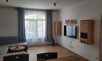 A modern living room with a gray sofa and a round table. Large windows let in plenty of light, and there is a stylish wall with a TV and wooden shelves.