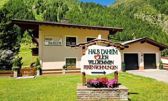 A cozy holiday home in the mountains with a green lawn. A welcome sign in the foreground says "Haus Daheim".