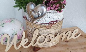 A decorative flower pot with colorful plants and a heart made of metal. Next to it lies a handmade wooden "Welcome" sign.