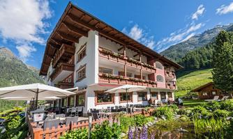 A traditional hotel in the mountains with a beautiful terrace and garden.  
Surrounded by green meadows and majestic mountains under a clear sky.