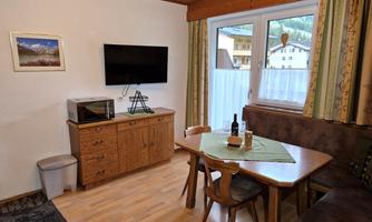 A cozy living space with a dining table and chairs. There is a television on the wall, and the room has large windows with a view outside.
