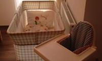 A crib and a high chair are in a bright room. The bed is decorated with a blanket, and the windows let in a lot of light.