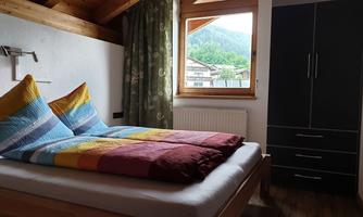 A cozy bedroom with a large window and a view of the mountains. The bed is colorfully decorated and the room has wooden details.