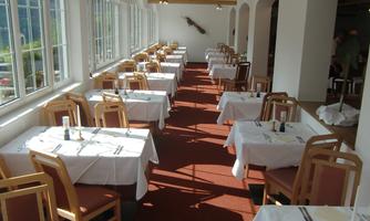 A bright restaurant with many tables covered with white tablecloths.  
Large windows provide natural light and a pleasant atmosphere.