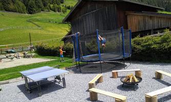 A beautiful outdoor area with a trampoline and a ping pong table. In the background, there is a wooden house and a green landscape.