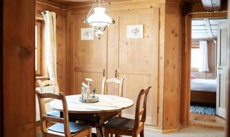 A cozy dining area with a round wooden table and four chairs. The walls are made of wood, and there is a warm light from a hanging lamp.