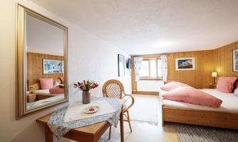A cozy room with wooden paneling, a double bed, and a small dining table. A mirror and windows provide bright lighting and an inviting atmosphere.