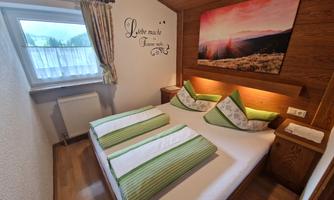 A cozy bedroom with a double bed and green pillows. A beautiful landscape greeting hangs on the wall, and a window lets in a lot of daylight.