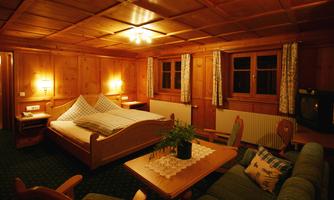 A cozy bedroom with wood paneling and two windows. There is a large bed, a table, and a comfortable armchair.