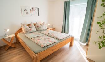 A bright bedroom with a large wooden bed and floral bedding. Green curtains hang in front of the window, and there is a small lamp on a nightstand.