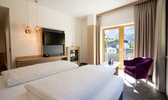 A modern hotel room with a large bed and a television. The large window front offers a view of the mountains and lets in plenty of daylight.