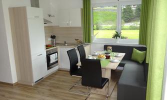 A modern kitchen with a dining table and black chairs. Large windows let in plenty of daylight and the room is designed in light colors.