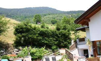 A beautiful garden with loungers and plenty of greenery lies in front of a bright house. In the background, gentle hills and forests stretch out.