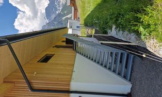 A modern building with wooden cladding and a metal staircase. In the background, there are green meadows and mountains visible in nice weather.