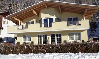 A modern house with a large balcony and a light yellow facade. The surroundings are snow-covered, surrounded by snow-covered trees.