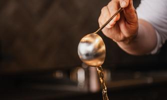 A hand pours a clear broth into a bowl. Dark lamps hang above the bowl, illuminating the room.