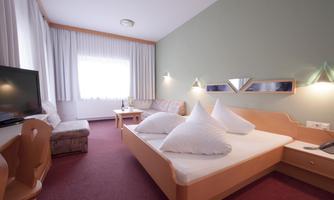 A bright room with a double bed, two large pillows, and a cozy sofa. The room is equipped with a television and a desk, and the window lets in plenty of daylight.