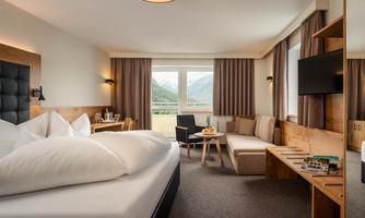 A modern hotel room with a large bed and a cozy seating area. Large windows offer a view of the surrounding mountains.