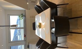 A modern dining area with a white table and six black chairs. In the background, there are windows with a view of a hilly landscape.