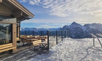 A terrace with wooden furniture and a view of snow-covered mountains. The sky is clear and the landscape is majestic.
