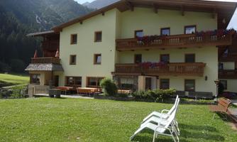 A cozy hotel with balconies and flowers on a meadow. In the foreground, there are loungers and the mountains are visible in the background.