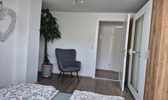 A modern room with two beds and a cozy armchair. A plant stands in the corner, and a door leads outside.