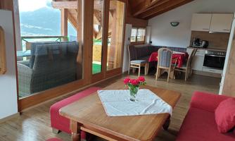 A cozy accommodation with a wooden ceiling and large windows. The room is furnished with a table, red sofas, and a modern kitchenette.