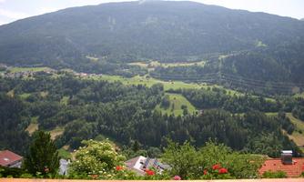 A picturesque mountain landscape with green meadows and wooded slopes. In the foreground, some houses and flowers can be seen.