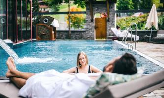 A relaxing pool area with a woman in the water and a man lying on a lounger. Surrounded by green trees and modern design.