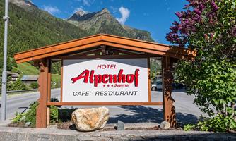 A sign of the Alpenhof hotel with the inscription "Café - Restaurant". In the background, green mountains and a blue sky can be seen.