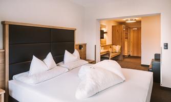 A modern bedroom with a large bed and comfortable pillows. The decor is bright and inviting, with wooden details and an additional living area.