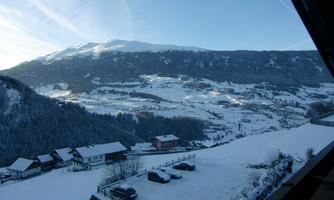 A winter mountain landscape with snow-covered hills. In the foreground, some houses and parked cars can be seen.