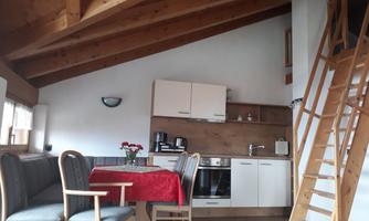 A cozy living space with wooden beams, a kitchen, and a dining table. A staircase leads to an upper area.
